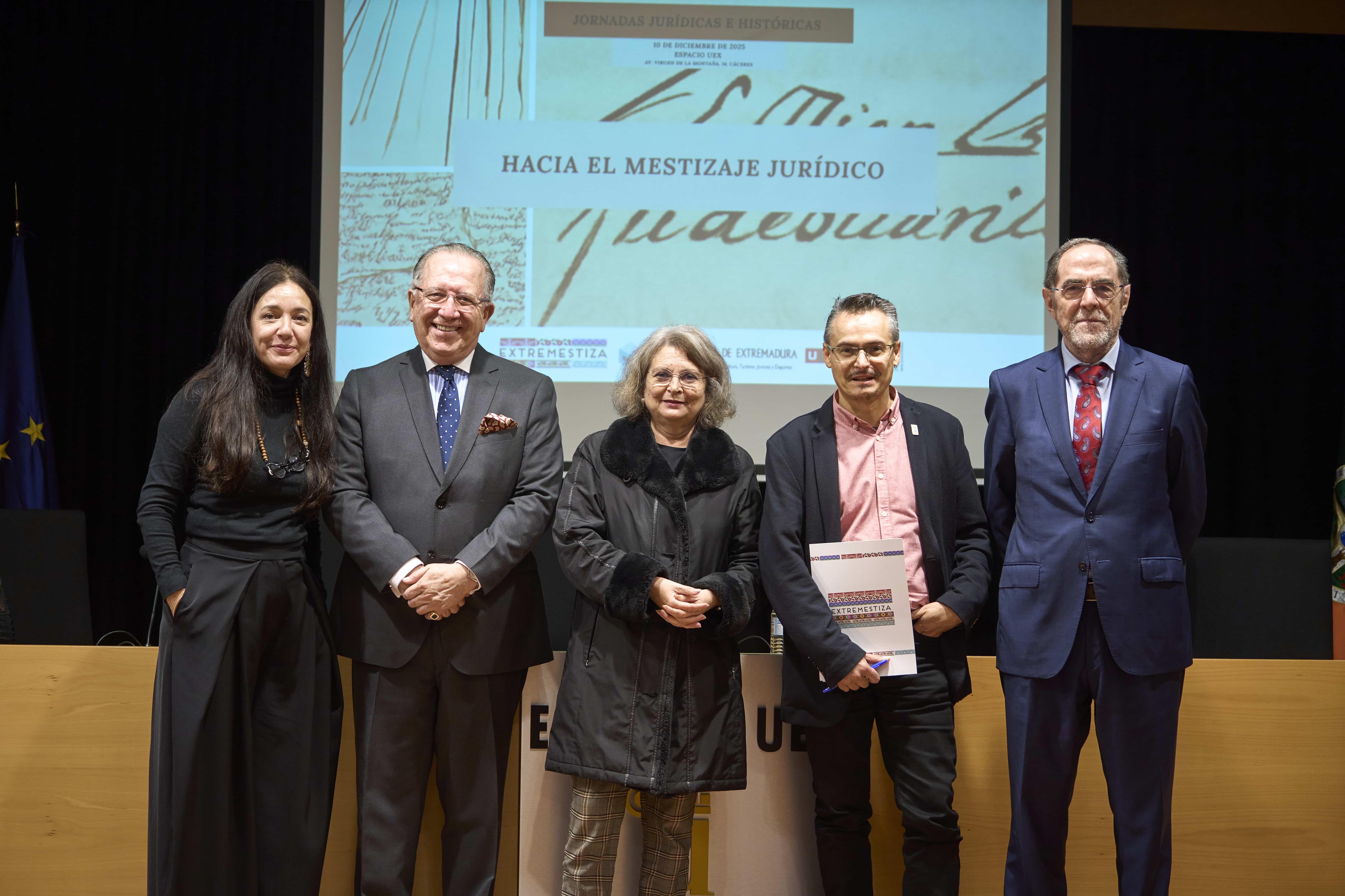 Five adults in formal attire stand on a stage posing for a photo, with a projected banner behind them reading 'HACIA EL MESTIZAJE JURÍDICO'.