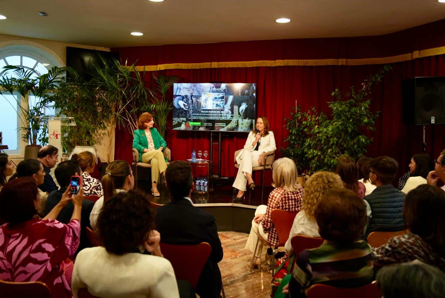 Two women sit on stage in a panel discussion, with a large screen displaying slides behind them and an attentive audience in a conference room with red curtains and potted plants.