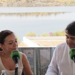 Two radio hosts interview outdoors by a lake, speaking into green microphones with notes on the table and a banner behind them reading EXTREMESTIZA Junta de Extremadura.