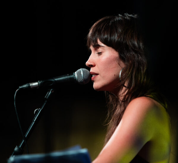 Female singer in profile singing into a microphone on a dimly lit stage with a dark background, eyes closed.