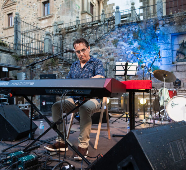 Musician playing a Yamaha MOXF8 keyboard on an outdoor stage with drums and a stone wall behind him.