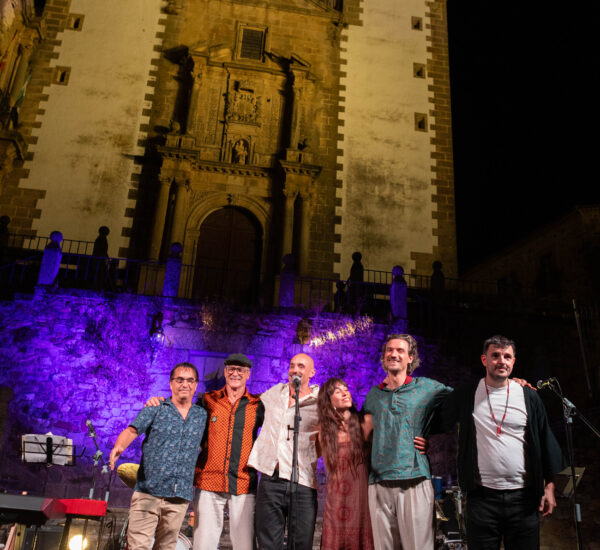 Group of musicians and singers posing on a lit stage at night, with a historic church façade and twin towers behind them.