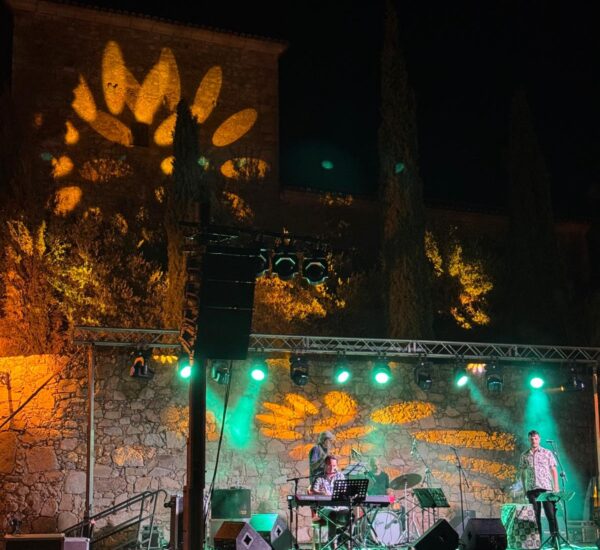 Nighttime outdoor concert on a stone-stage with musicians under green lights and a sunflower-shaped projection on the wall behind them.