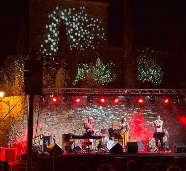 Nighttime outdoor concert with a band performing on a stone stage and red lights, backdrop lit by bubble-like projections on the wall.